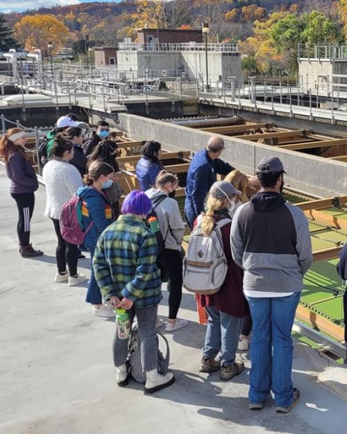 		Students standing around an open wastewater treatment facility, which looks like a big cement rectangle with green water and wooden separaters.
	