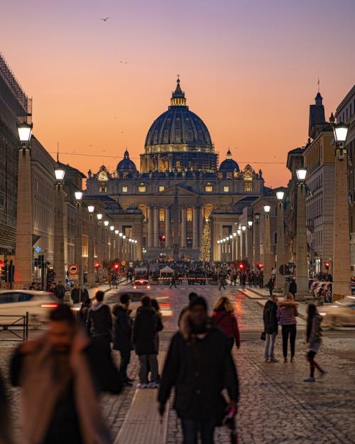		Dark, late evening sky in purple and orange over the ornate dome of St. Peter's Church in Rome; many pedestrians crowd cobblestone sidewalks in the foreground
	