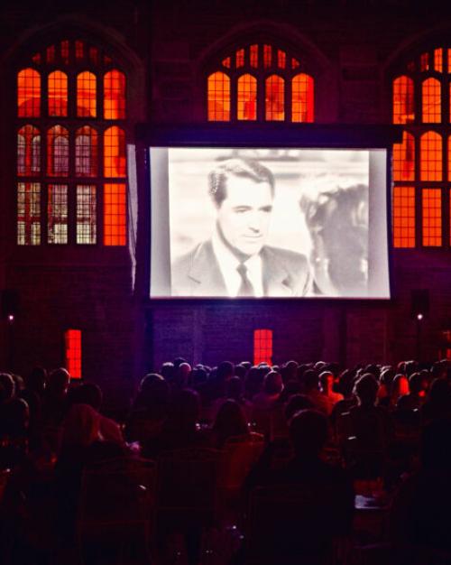 		Movie screen outdoors, showing a black and white still of Jimmy Stewart, with red-lit windows behind it. 
	