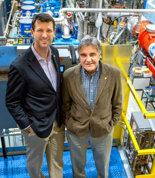  Two physicsists stand in front of accelerator equipment