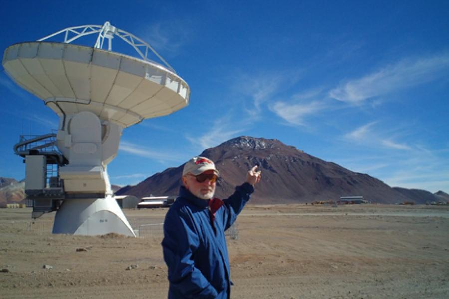 Riccardo Giovanelli in blue jacket, baseball cap, sunglasses and white beard, points to the top of a mountain in the desert. A telescope is seen in the background.