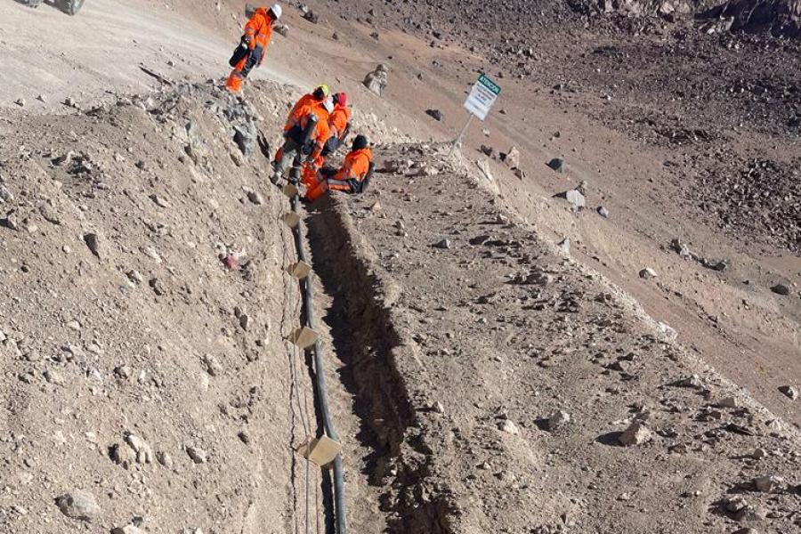 Construction workers in orange jumpsuits resting at one end of a long trench filled with three cables dug through a desert landscape.