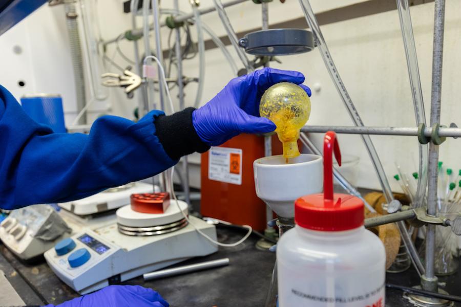 Arm and hand clad in dark blue lab clothing and gloves holds a glass bulb filled with viscous yellow liquid