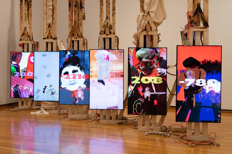 Art work consisting of six vertical screens showing bright digital images, set on wooden easels
