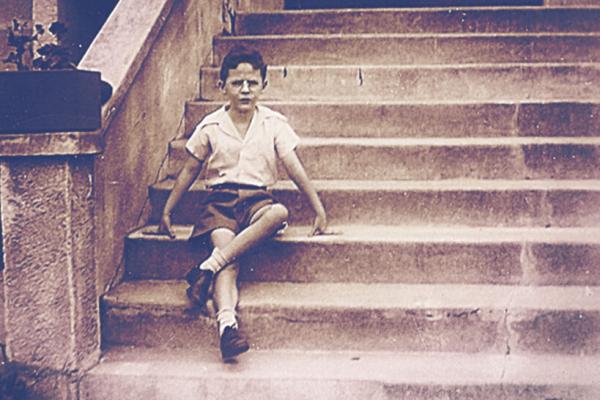 A boy seated on stone steps, dressed in shorts and a white shirt. Black and white historical image