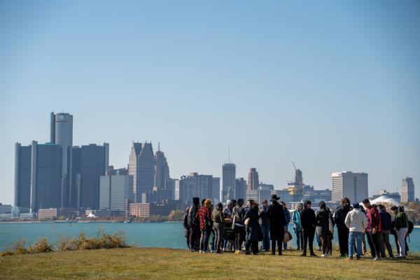 Several people stand on a grassy space looking over a river with a city on the other side