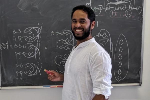 Darren Pereira in a white shirt rolled up to his elbows, smiling with a black beard and mustache, standing at chalkboard in front of diagrams.