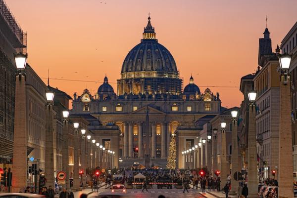 Dark, late evening sky in purple and orange over the ornate dome of St. Peter's Church in Rome; many pedestrians crowd cobblestone sidewalks in the foreground