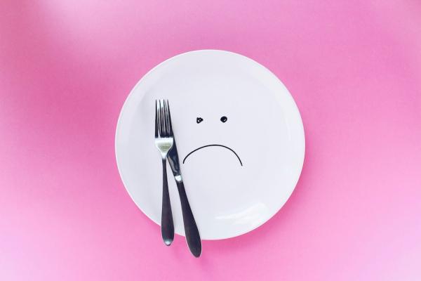 White plate on a pink background, with a fork and a knife. There is a sad face drawn on the plate