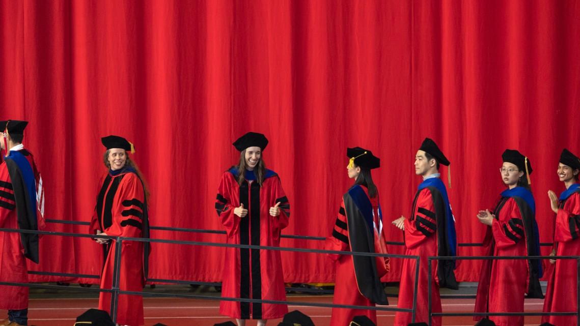 Several people in long red robes and black caps walk in a line against a red background; one turns to give a thumbs up