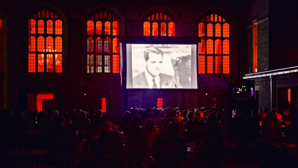 Movie screen outdoors, showing a black and white still of Jimmy Stewart, with red-lit windows behind it. 