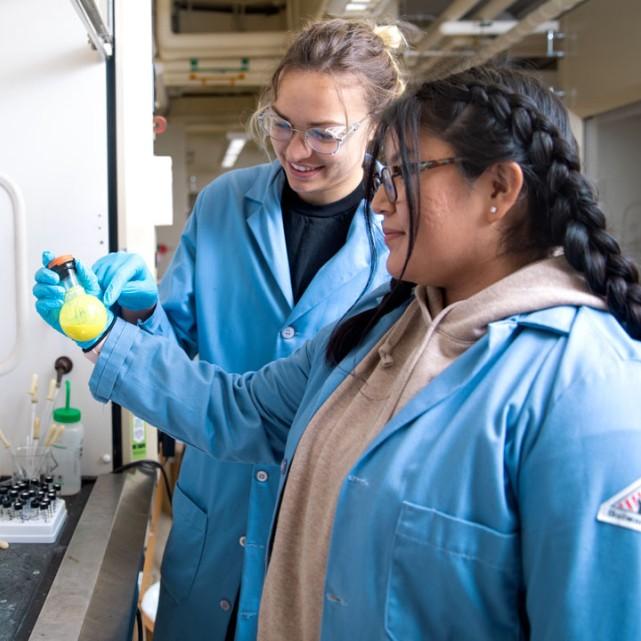 Two students wearing lab coats examining a beaker of something yellow in a lab.