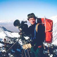 Person in hiking gear with a large camera; mountains in background