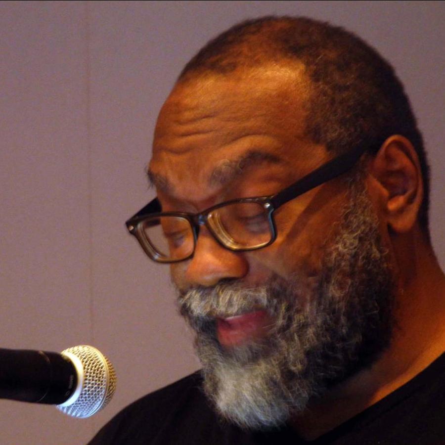 Fred Moten, with a mostly white beard and black glasses in front of a microphone