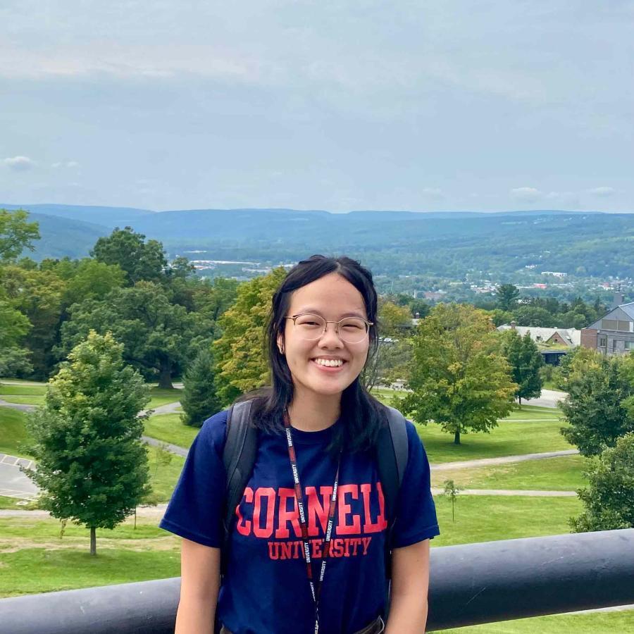 Eunice Ngai in a blue Cornell t-shirt with trees and the Ithaca hills in the background.