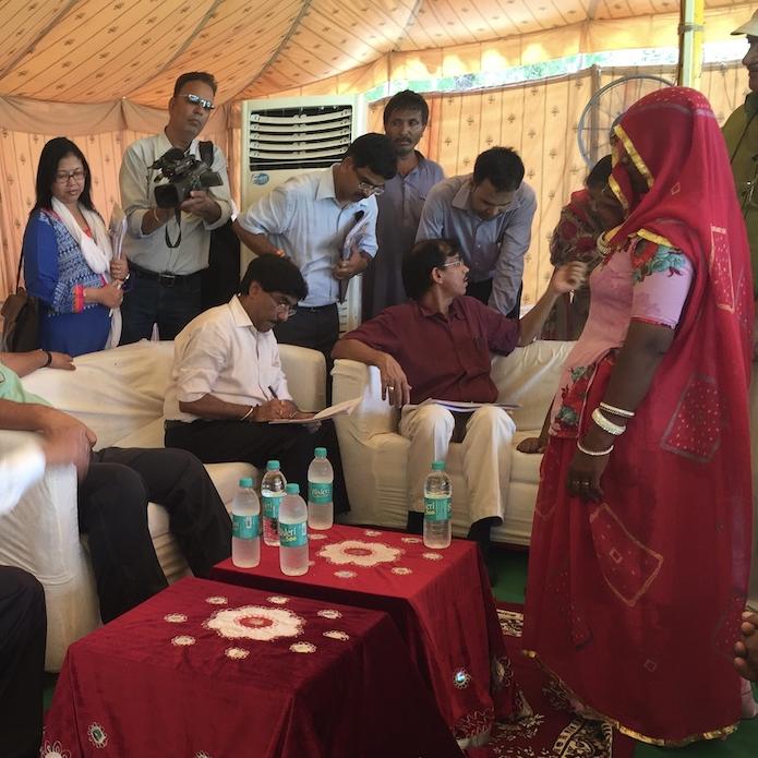 Among several people in a tent, a woman in red traditional clothing faces three officials in white shirts and black trousers, sitting on a couch