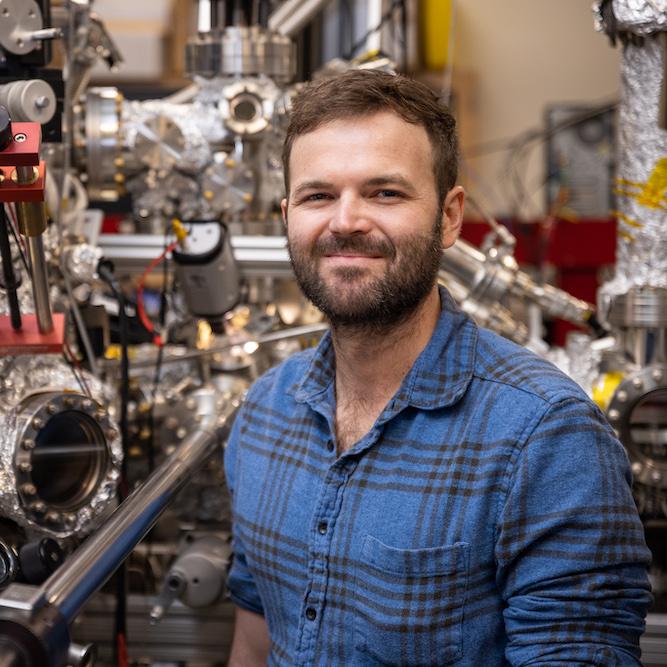 Person in blue shirt stands in front of complicated silver equipment