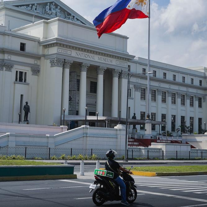Motorcycle drives past a stone "National Museum" fronted by the Philippine flag