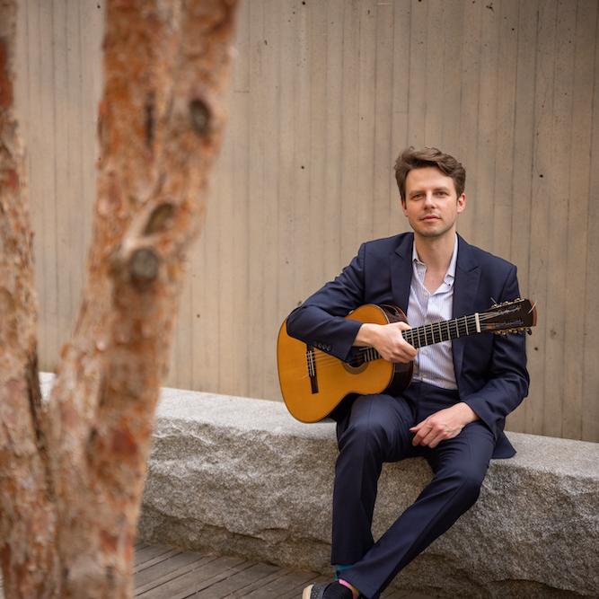 Person sitting on a stone wall, holding a guitar near trees