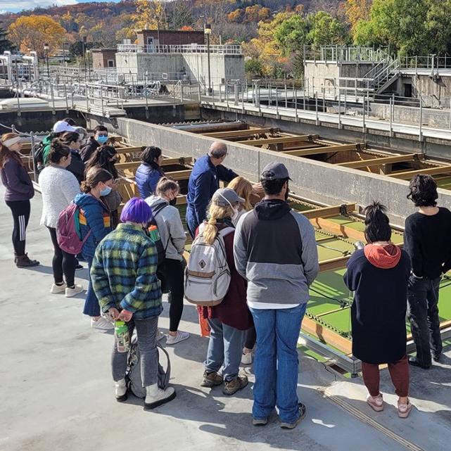 Students standing around an open wastewater treatment facility, which looks like a big cement rectangle with green water and wooden separaters.