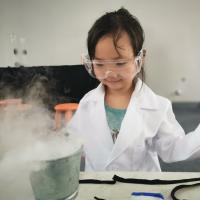 Small person wearing safety goggles and a white lab coat, smiling as smoke pours out of a beaker on a lab bench