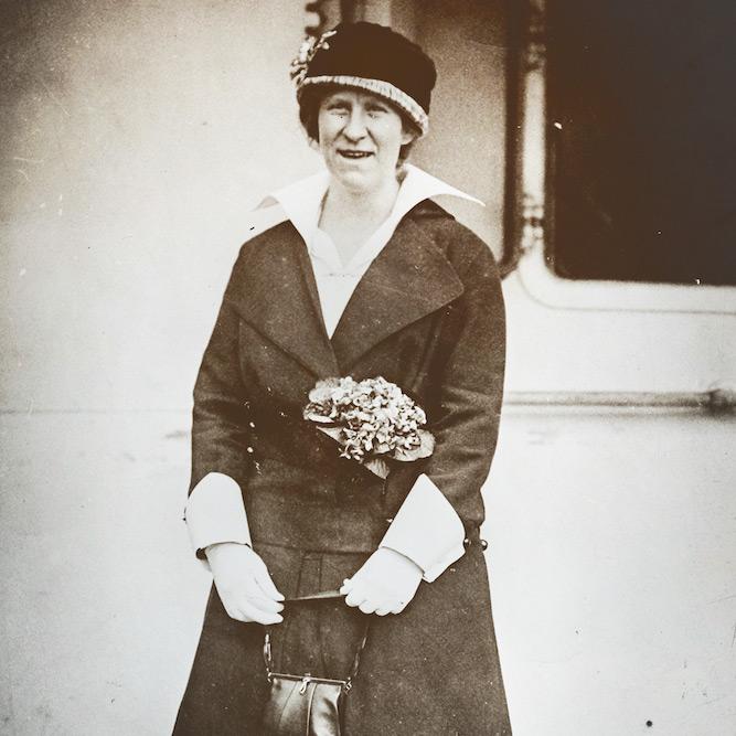 Black and white photo from 1914: a woman in a dark suit and hat highlighted by flowers stands on a wooden dock