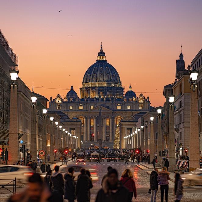 		Dark, late evening sky in purple and orange over the ornate dome of St. Peter's Church in Rome; many pedestrians crowd cobblestone sidewalks in the foreground
	