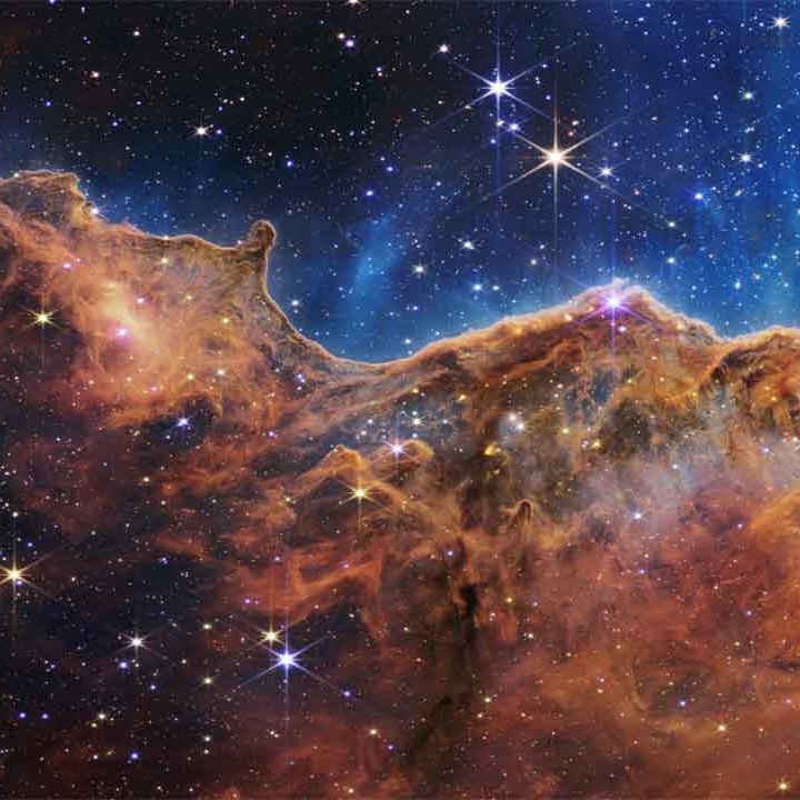 A field of stars in the background and in the foreground a colorful cliff-shaped mass of cosmic gases.