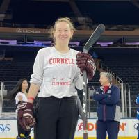Person wearing hockey gloves and a Cornell t-shirt, carrying a hockey stick and smiling