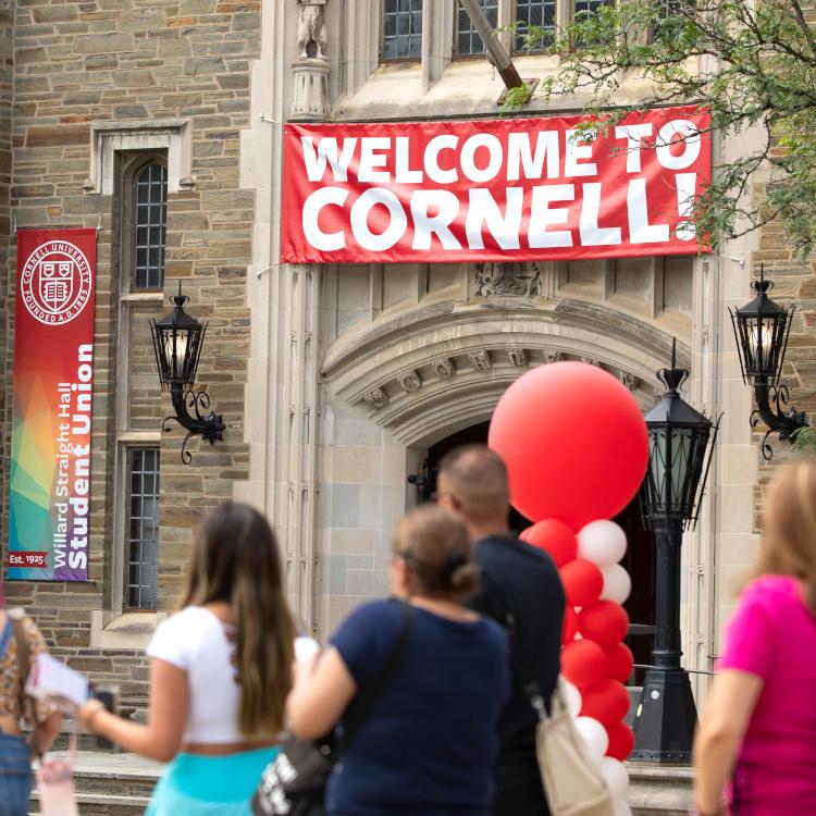 		Several people walk past a building with a red and white banner that says "Welcome to Cornell." There are red balloons
	