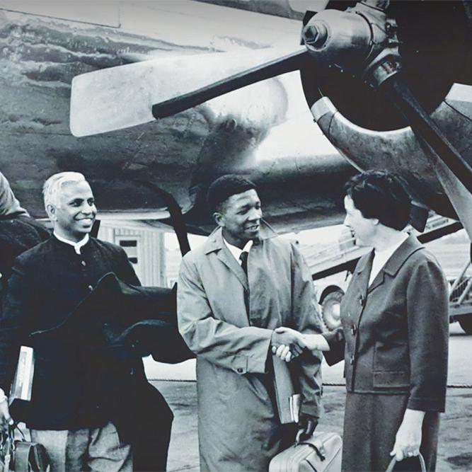 		Black and white historic photo: Five people getting off a plane; one is shaking hands with a person wearing a suit at the bottom of the stairacase
	