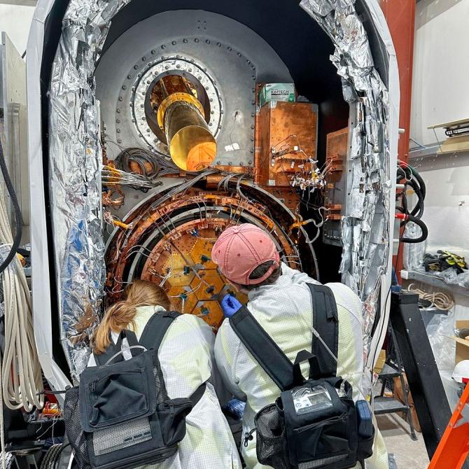 the backs of two people who are facing a large opening containing a scientific instrument, to work on it. 