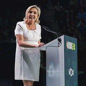 		Marine Le Pen in a short white dress facing the audience, standing at a podium that says "Viva24"
	