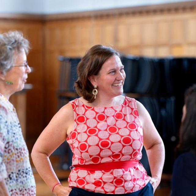 		Person in red shirt smiles, hands on hips, while speaking with a circle of other people
	