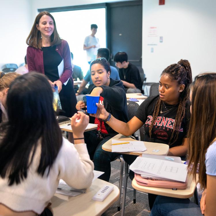 		About six students sit in desks and interact using a blue Solo cup while an instructor stands by
	