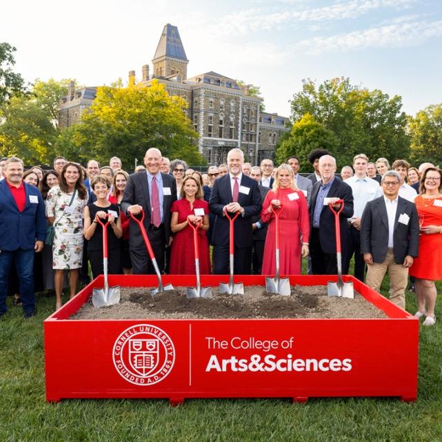 		A crowd of about 75 people stands behind a low box full of dirt; six people in the front hold shovels with red handles during a ceremonial "groundbreaking" event
	