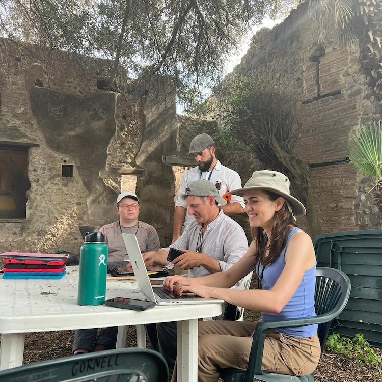 		Four people work at a plastic patio table in the midst of ancient ruins: they are archaeologists on an excavation site
	