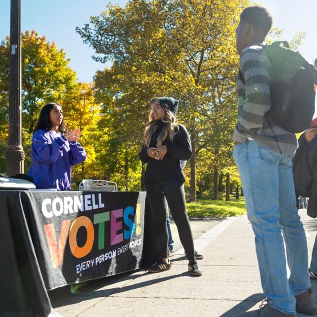 		People with backpacks and jeans stand in front of a table, set outdoors, that's labeled "Cornell VOTES"
	