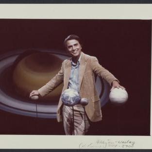 		Carl Sagan in casual jacket, smiling, with planet models swirling around him and his hands on top of two different planets
	