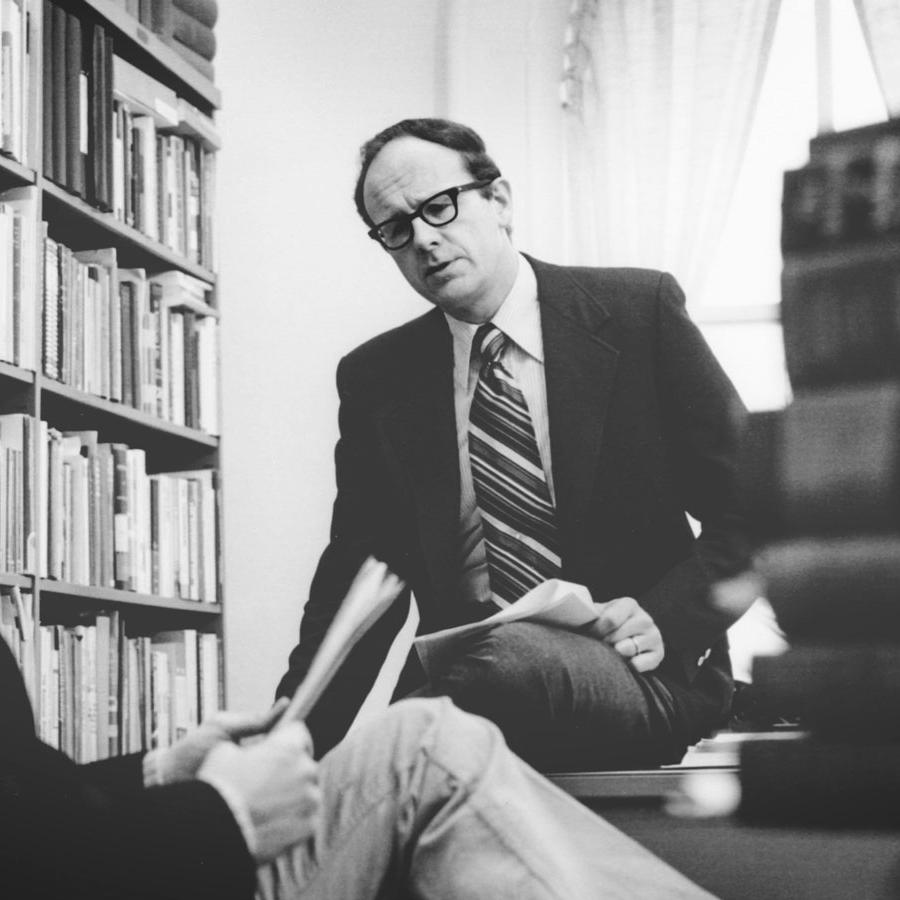 Walter LaFeber in suit and striped tie in his office with bookshelf on one side and a pile of books on the other, talking to someone.