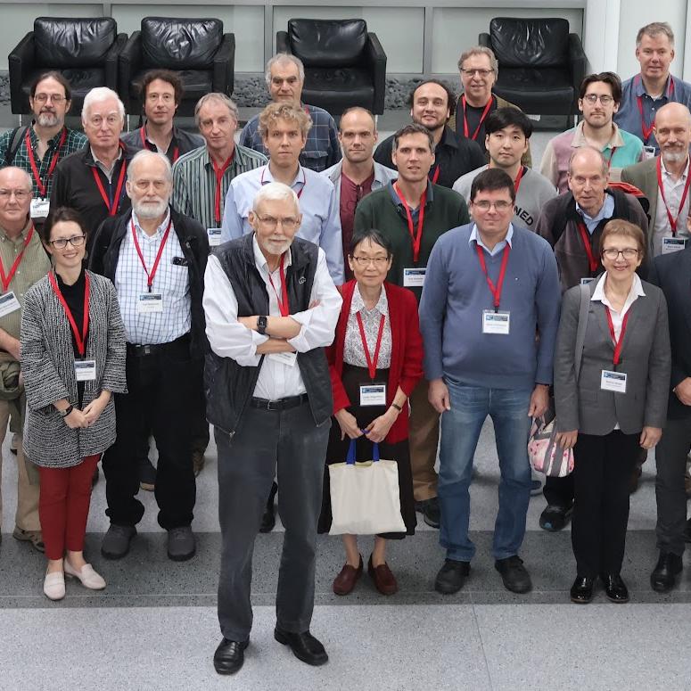 Peter Lepage, wearing a black vest, stands in front of a large group of people assembled to talk about physics and honor his work