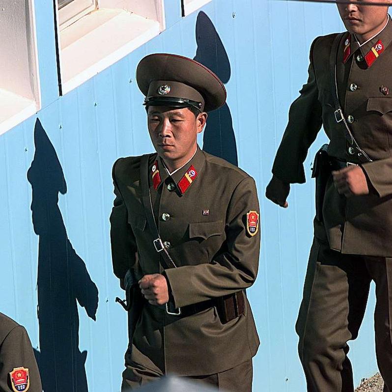 Soldiers in brown formal uniforms march in line beside a blue wall