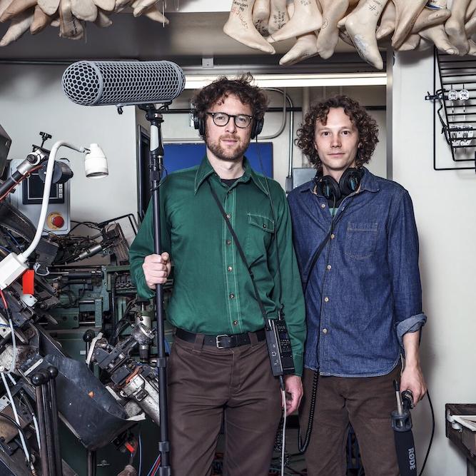 Two people in casual clothes stand in a room full of bric-a-brac, holding professional grade recording equipment