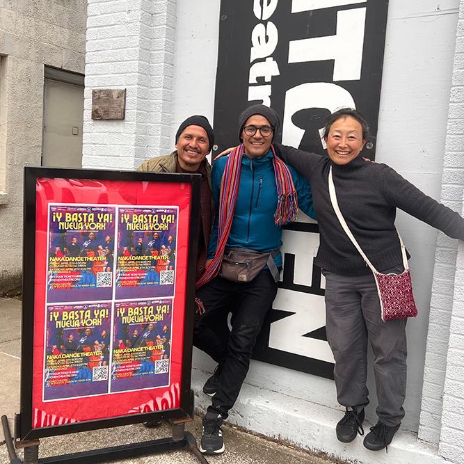 		Three people stand near a red production poster outside a theater
	