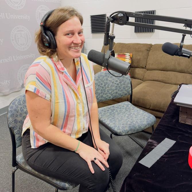 Alexis Boyce, wearing headphones in front of a big microphone, smiles at the camera, wearing a bright colored, striped shirt.