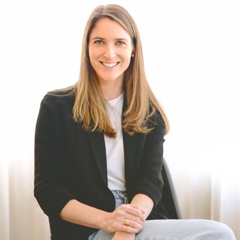 		Jenna Barnes, dressed in jeans, open sweater and t-shirt, with long brown hair, seated and smiling.
	