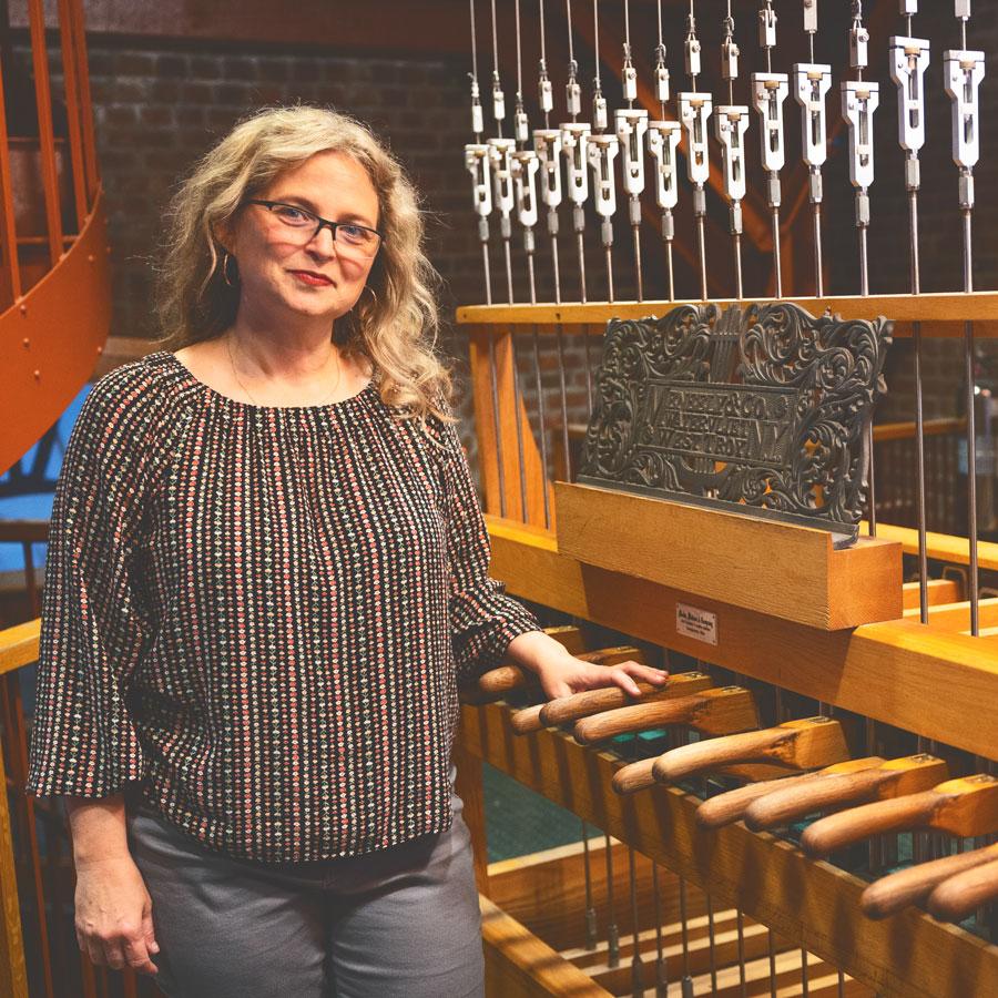 Jennifer Lory-Moran, in multi-colored stripped shirt and long blond hair, is smiling while standing next to the controls for the Cornell chimes up in McGraw Tower.