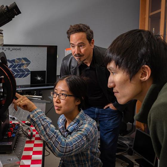 		Three people look intensely at a small black and red machine in a science laboratory
	