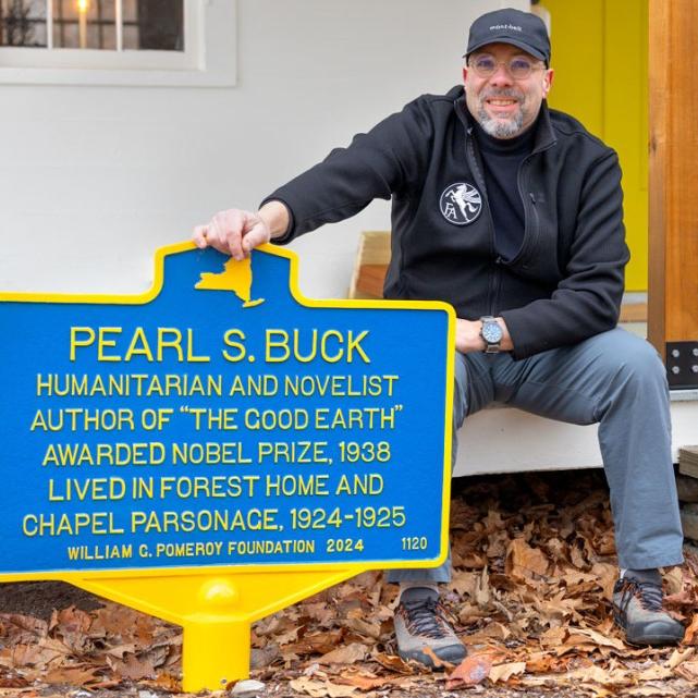 		Person sits on a porch with one hand on a blue historic marker
	