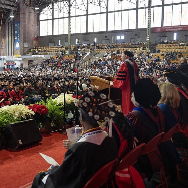 Large indoor gymnasium (Cornell's Barton Hall) decorated with pointsettias and filled with people wearing caps and gowns